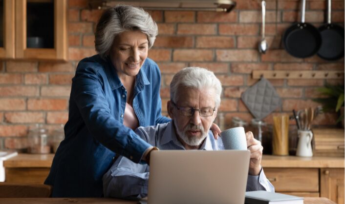 Elderly couple in a cozy kitchen, engaged at a laptop. Woman in denim shirt smiles, pointing to the screen, while man in glasses holds a mug, focused.