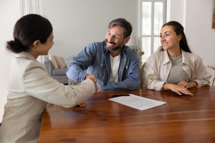Happy husband and wife shaking hands with real estate insurance agent, giving handshake to financial adviser over paper contract, smiling, laughing, closing house buying agreement