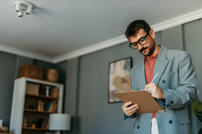 Professional man wearing a blazer and glasses writing on a clipboard, standing indoors